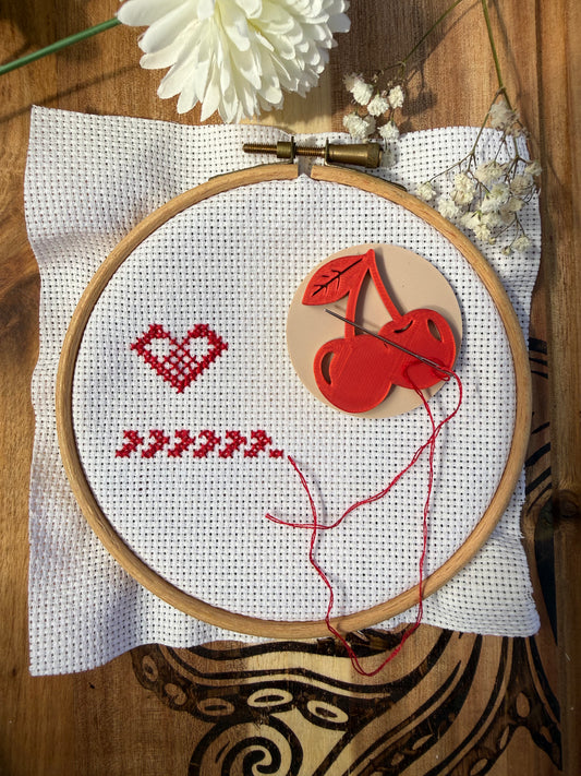 A wooden embroidery hoop holding white A4 fabric with a red stitched heart and chevron pattern, alongside a red cherry magnetic needle minder and thread. The hoop is placed on a white cloth over a dark wood surface, octopus with dried flowers and a white flower in the background.