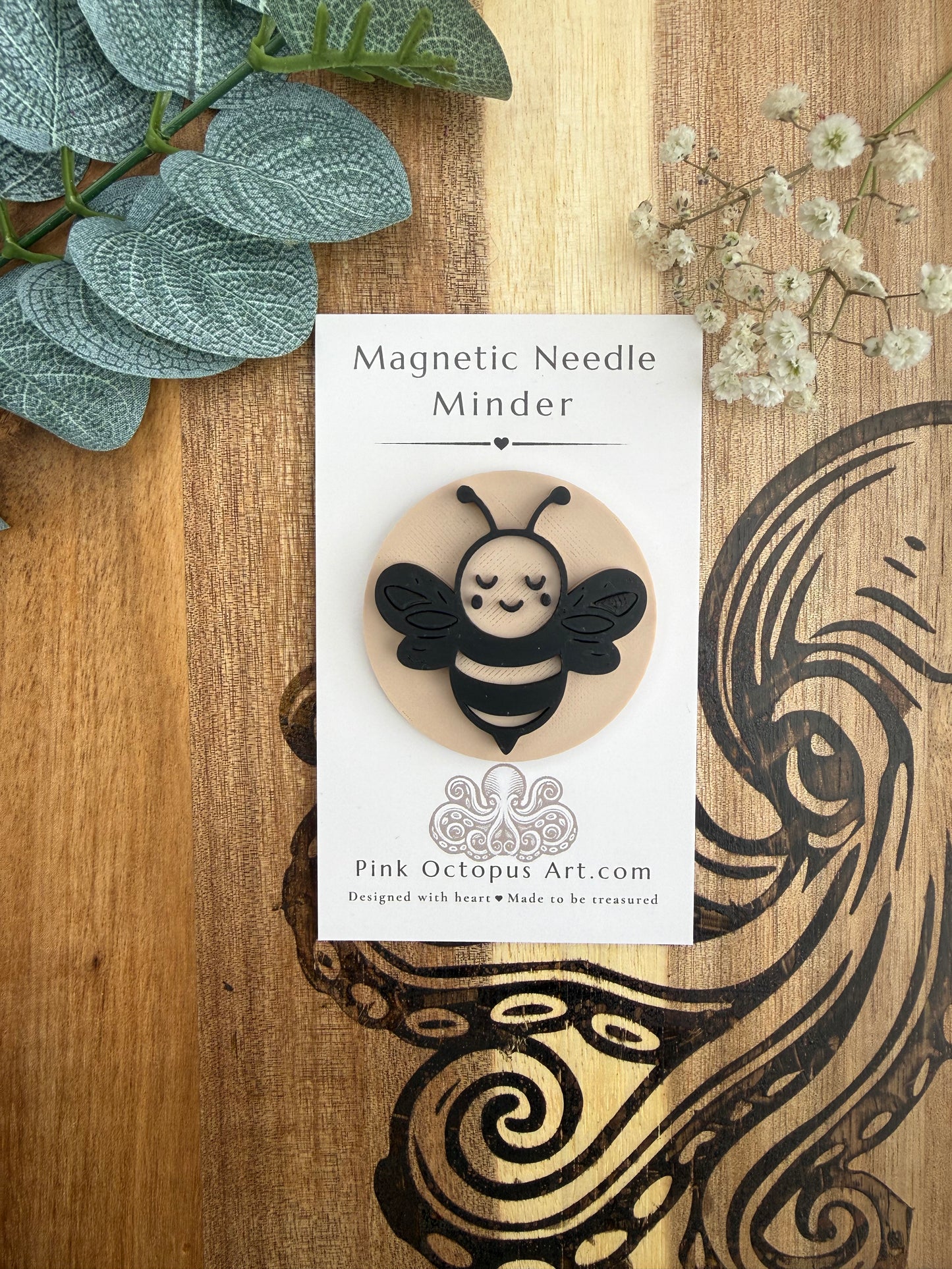 A decorative card displaying a bee-shaped magnetic needle holder labeled 'Magnetic Needle Minder' and 'Pink Octopus Art.com,' styled on a wooden surface with green leaves and small white flowers.