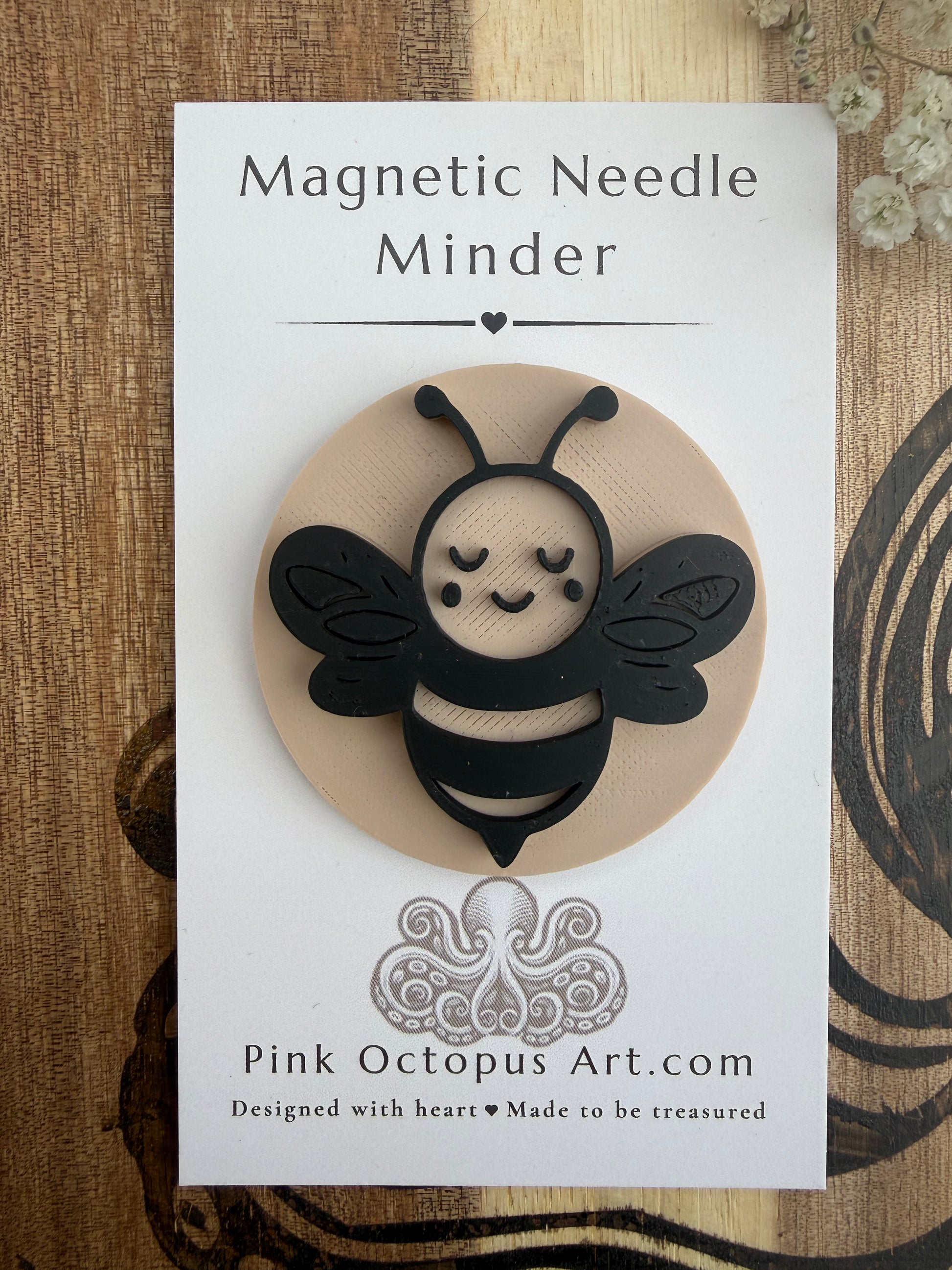A cute black and beige magnetic bee ornament mounted on a white backing card labeled 'Magnetic Needle Minder' with 'Pink Octopus Art.com' printed below, photographed on a wood surface.