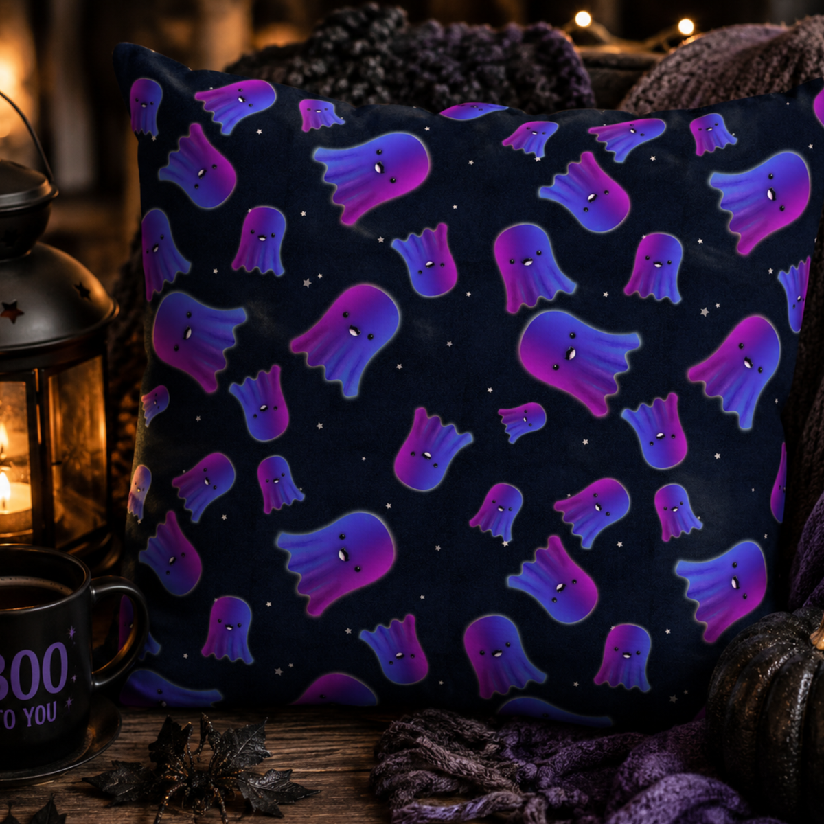 A square throw pillow with a dark navy blue background and a repeating pattern of purple and blue ghosts, styled in a cozy autumn setting with a lantern, mug, and knit blankets.
